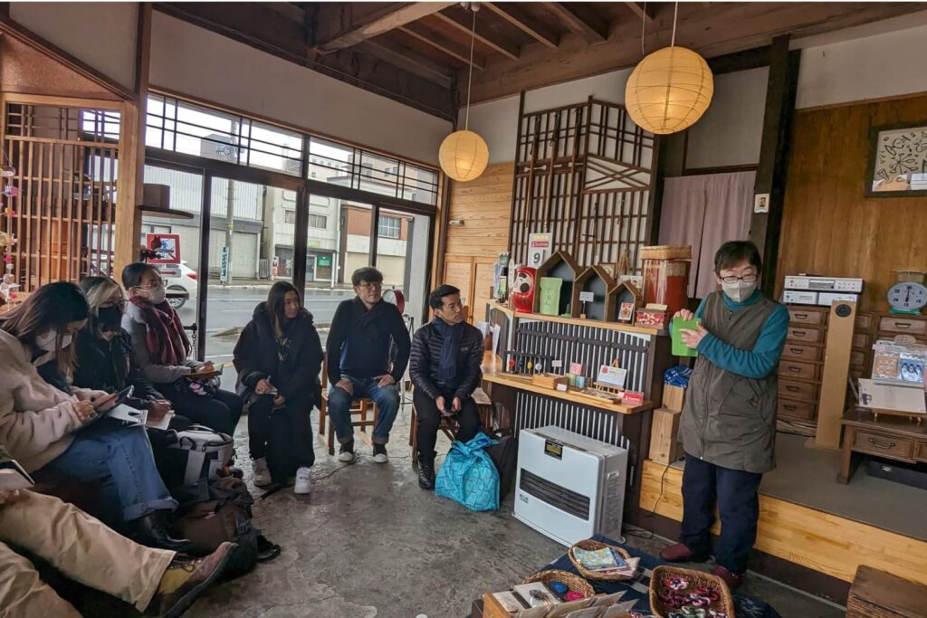 日本の伝統的な木造建物の室内で、複数の人々が座って話を聞いている場面。部屋の一角で講師が、紙を持ちながら説明している。後方には木製の棚や民芸品が並べられ、窓からは外の通りが見える。